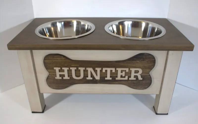Raised dog bowl stand with a light painted base, dark stained top, two stainless steel bowls and a bone-shaped nameplate reading Hunter in cream letters