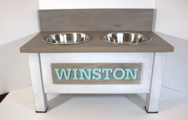 White dog bowl stand with a grey stained top and backsplash, two stainless steel bowls, and a grey nameplate reading Winston in light blue letters